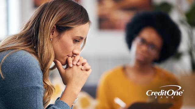 A young woman rests her chin on her hands as if she’s contemplating a crisis while another woman, who is perhaps her therapist or credit counselor, looks on with a clipboard.